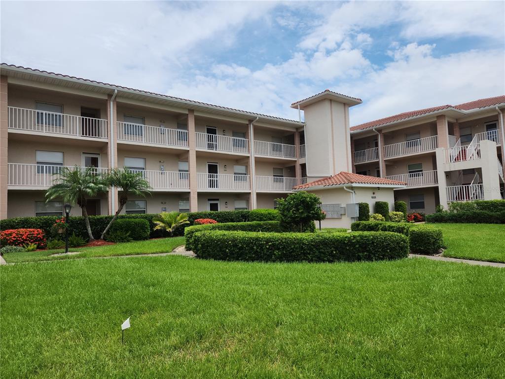 A three-story apartment building with a landscaped courtyard and a small tiled entrance structure — 6501 STONE RIVER ROAD UNIT 303, BRADENTON, FL