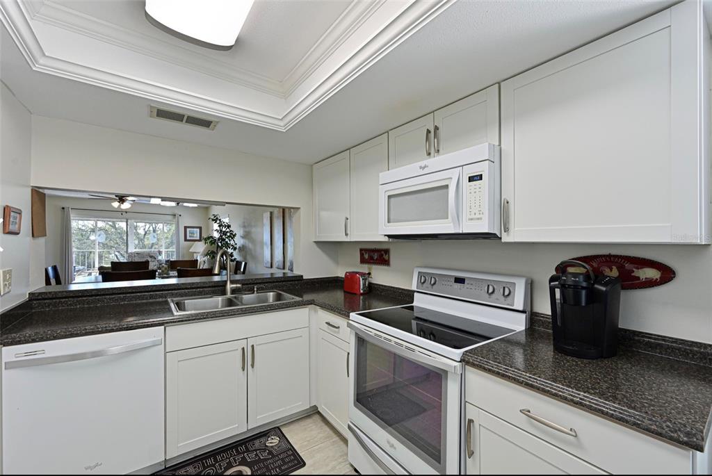 A kitchen features white cabinets, dark countertops, white appliances, and a pass-through window to a living area — 6501 STONE RIVER ROAD UNIT 303, BRADENTON, FL