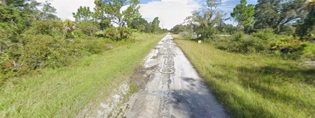 A narrow, weathered paved road stretches through a grassy, undeveloped lot with surrounding trees — Lot 68 CONSTITUTION DRIVE, NORTH PORT, FL