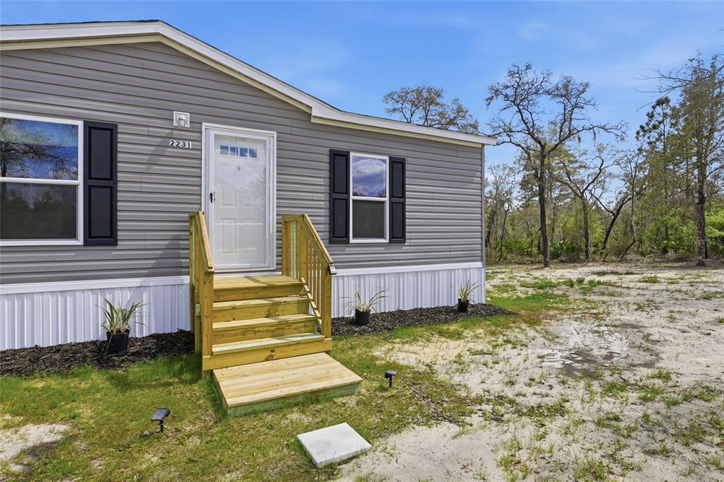 A gray manufactured home exterior features a white front door, wooden steps, and black shutters — 2231 SE 132ND COURT, MORRISTON, FL