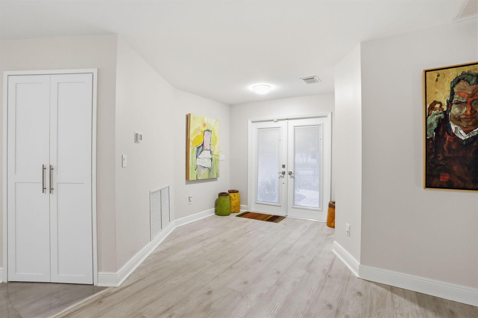 A bright entryway features light wood-look flooring, white walls, two paintings, and double glass doors — 2001 Embassy Drive, West Palm Beach FL 33401, WEST PALM BEACH, FL