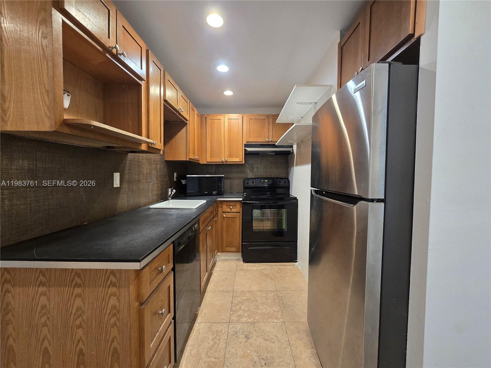 This galley kitchen features wood cabinets, black countertops, stainless steel appliances, and tile flooring — 223 S 17th Ave # 2, Hollywood FL 33020, HOLLYWOOD, FL