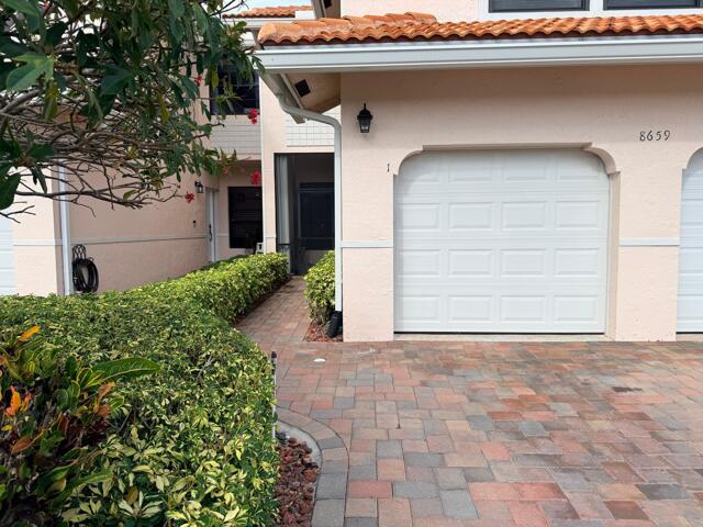 A beige townhouse exterior features a white garage door, a brick driveway, and a manicured hedge — 8659 Vía Reale # 1, Boca Raton FL 33496, BOCA RATON, FL