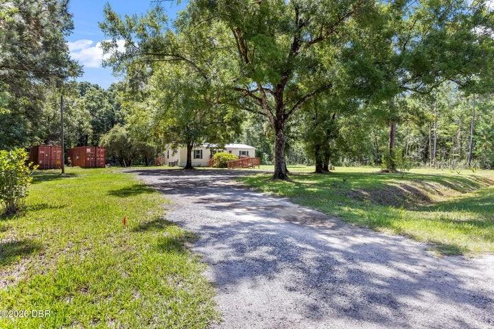 A gravel driveway leads to a single-story house surrounded by large trees and two containers — 10120 OLIVER AVENUE, HASTINGS, FL