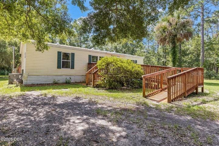 A light-colored mobile home features a wooden deck and wheelchair ramp in a wooded yard — 10120 OLIVER AVENUE, HASTINGS, FL