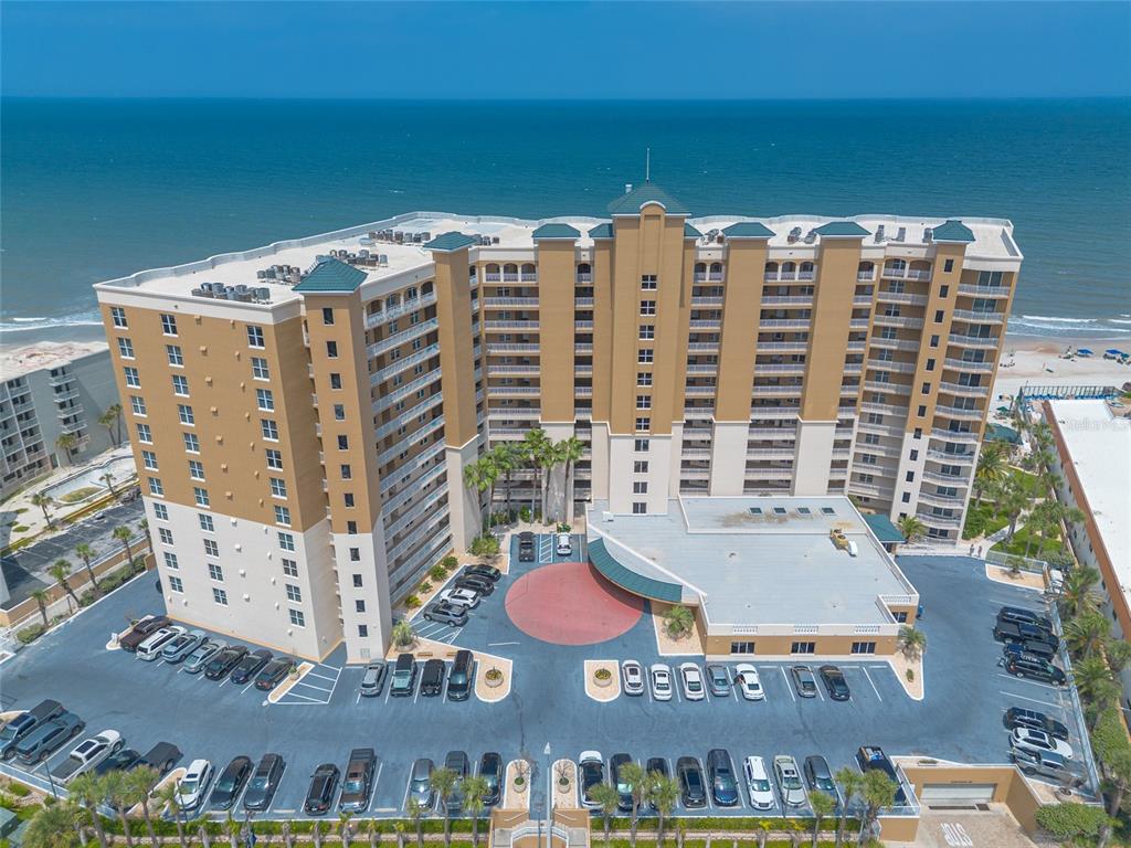 An aerial view shows a multi-story tan beachfront condominium building with a large parking lot — 2403 S ATLANTIC AVENUE UNIT 910, DAYTONA BEACH SHORES, FL