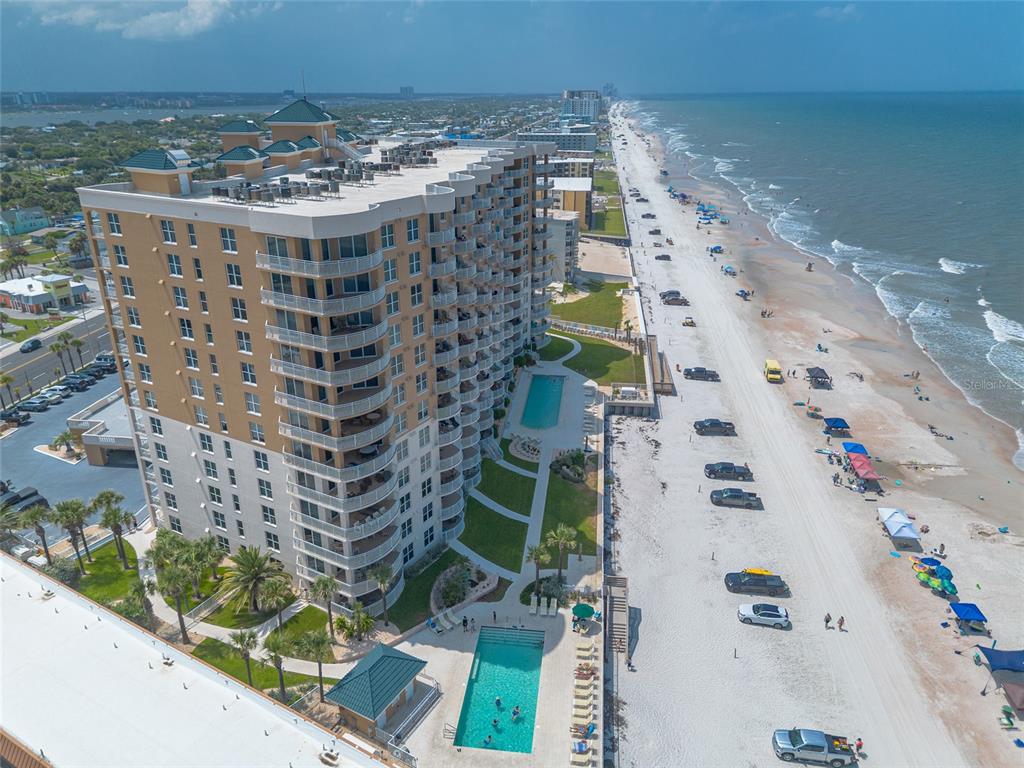 An aerial view shows a tall tan condominium building next to a sandy beach — 2403 S ATLANTIC AVENUE UNIT 910, DAYTONA BEACH SHORES, FL