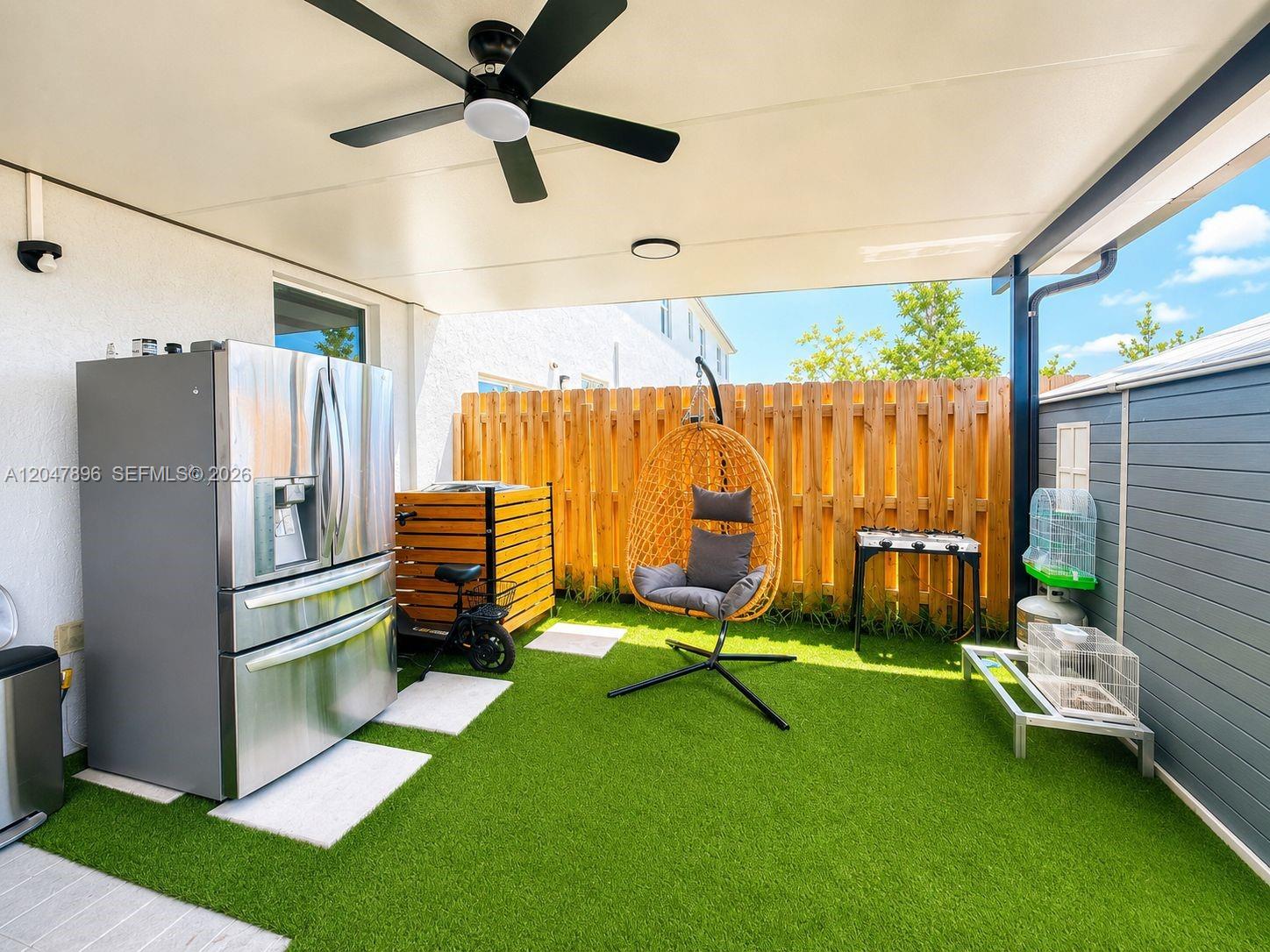 A covered patio features artificial turf, a stainless steel refrigerator, and a hanging egg chair — HOMESTEAD, FL