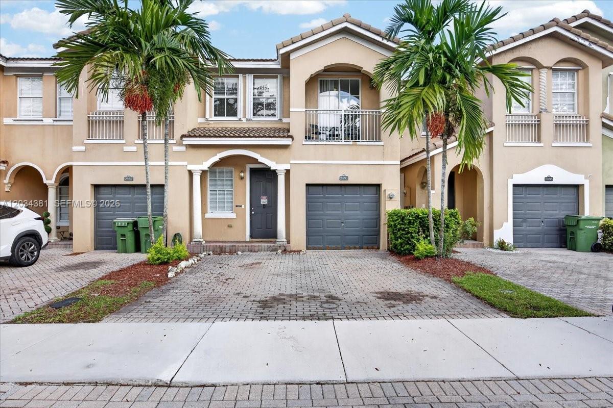 A row of three-story townhouses features beige stucco facades, gray garage doors, and palm trees — 2491 SE 13th Ct, Homestead FL 33035, HOMESTEAD, FL