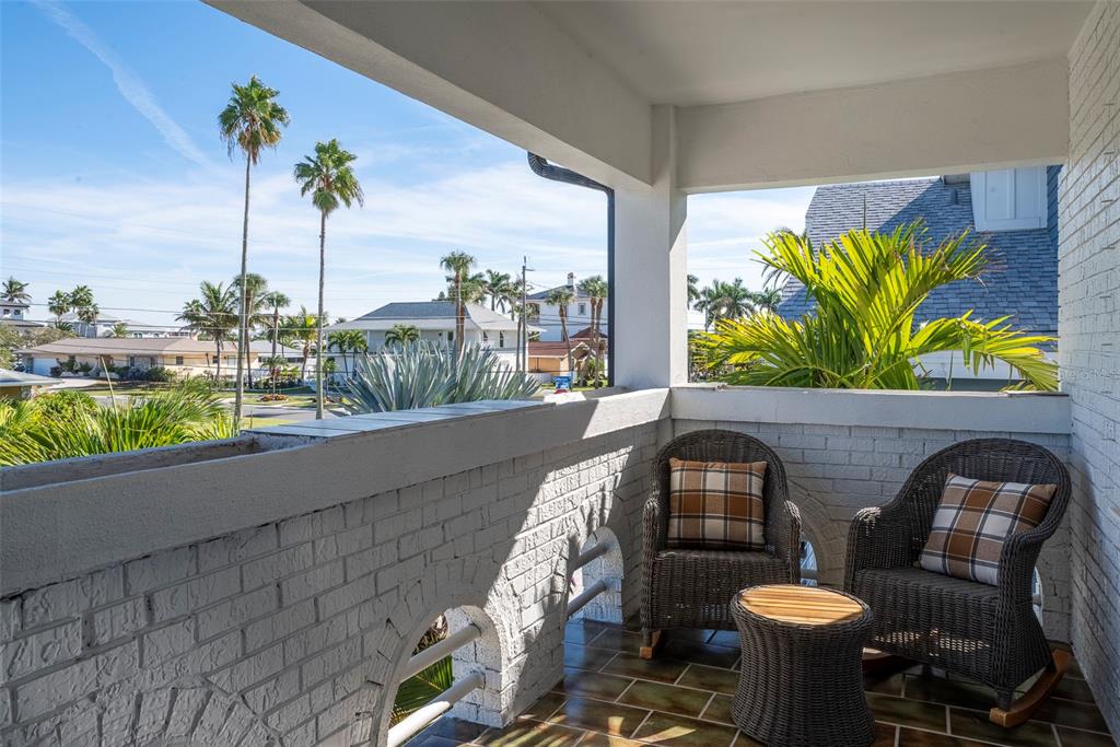 A covered balcony with white brick walls, wicker rocking chairs, and a view of palm trees — 2710 HIBISCUS DRIVE W, BELLEAIR BEACH, FL