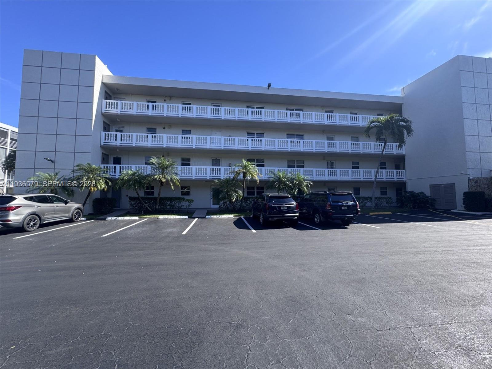 A three-story apartment building with white balconies sits behind a large paved parking lot — 1025 SE 3rd Ave # 102, Dania Beach FL 33004, DANIA BEACH, FL