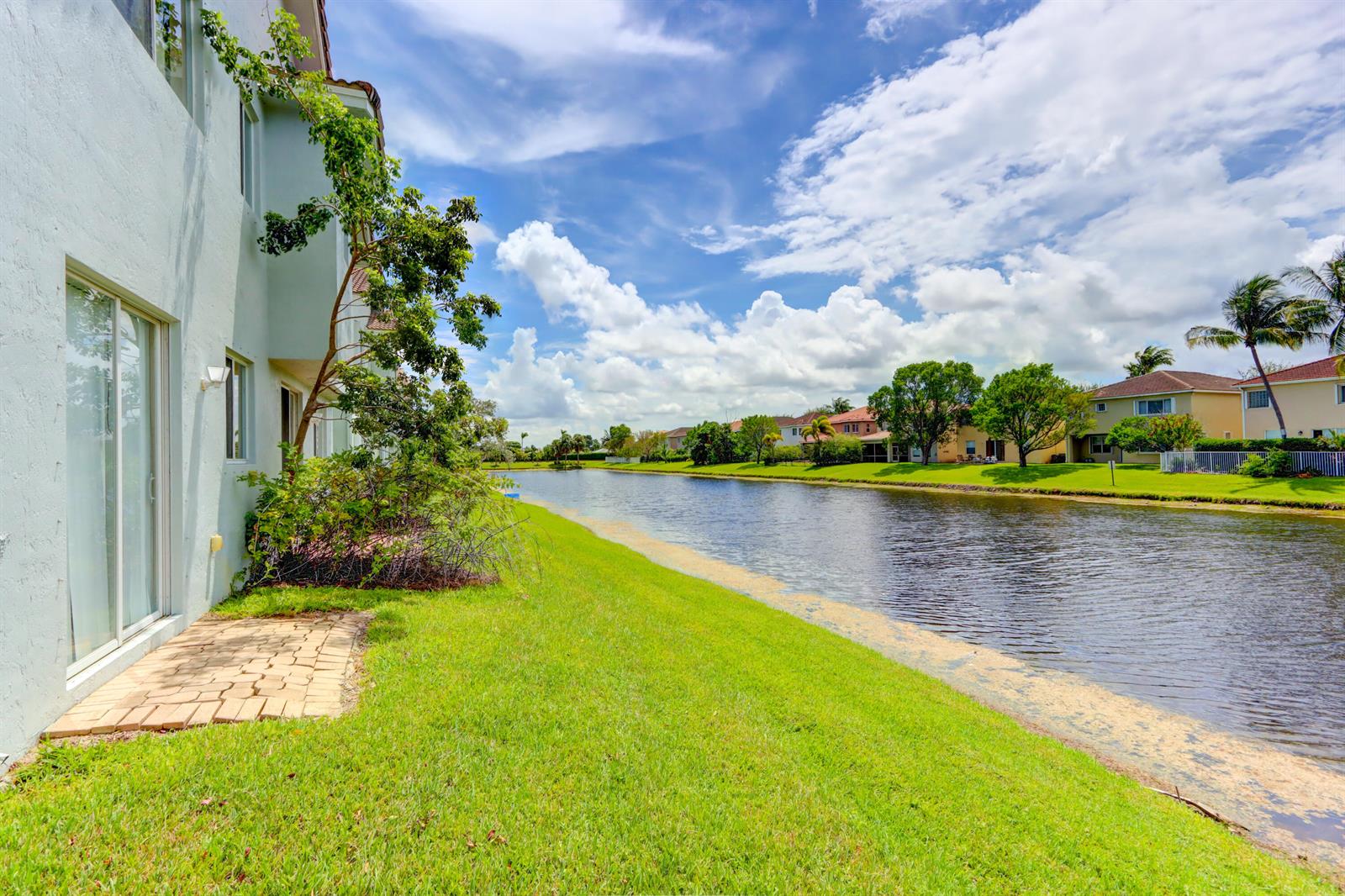 A view of a grassy backyard with a patio leading to a canal and houses — 3061 Laurel Ridge Circle, Riviera Beach FL 33404, RIVIERA BEACH, FL