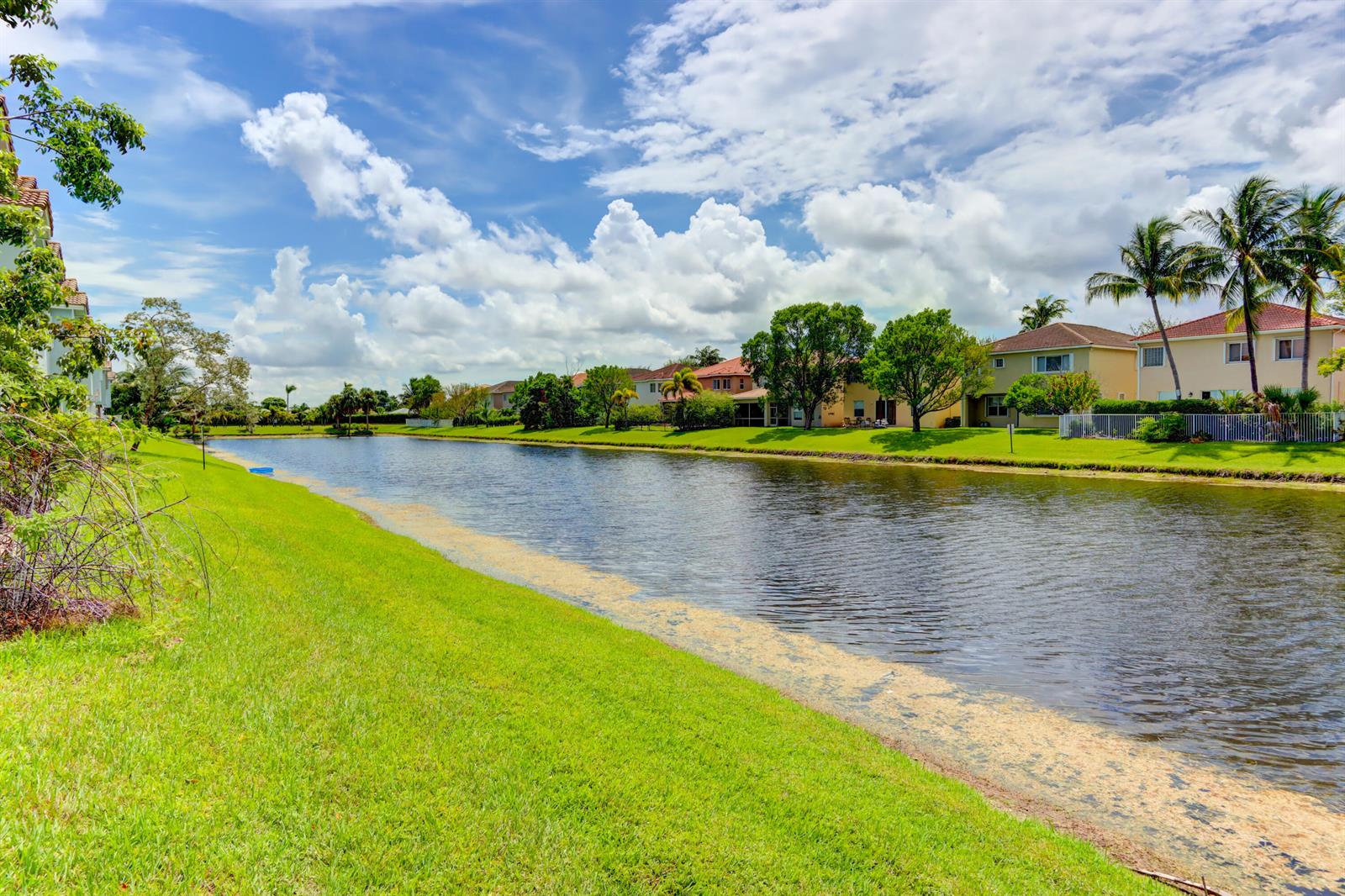 A grassy lawn borders a canal with residential houses and palm trees in the background — 3061 Laurel Ridge Circle, Riviera Beach FL 33404, RIVIERA BEACH, FL