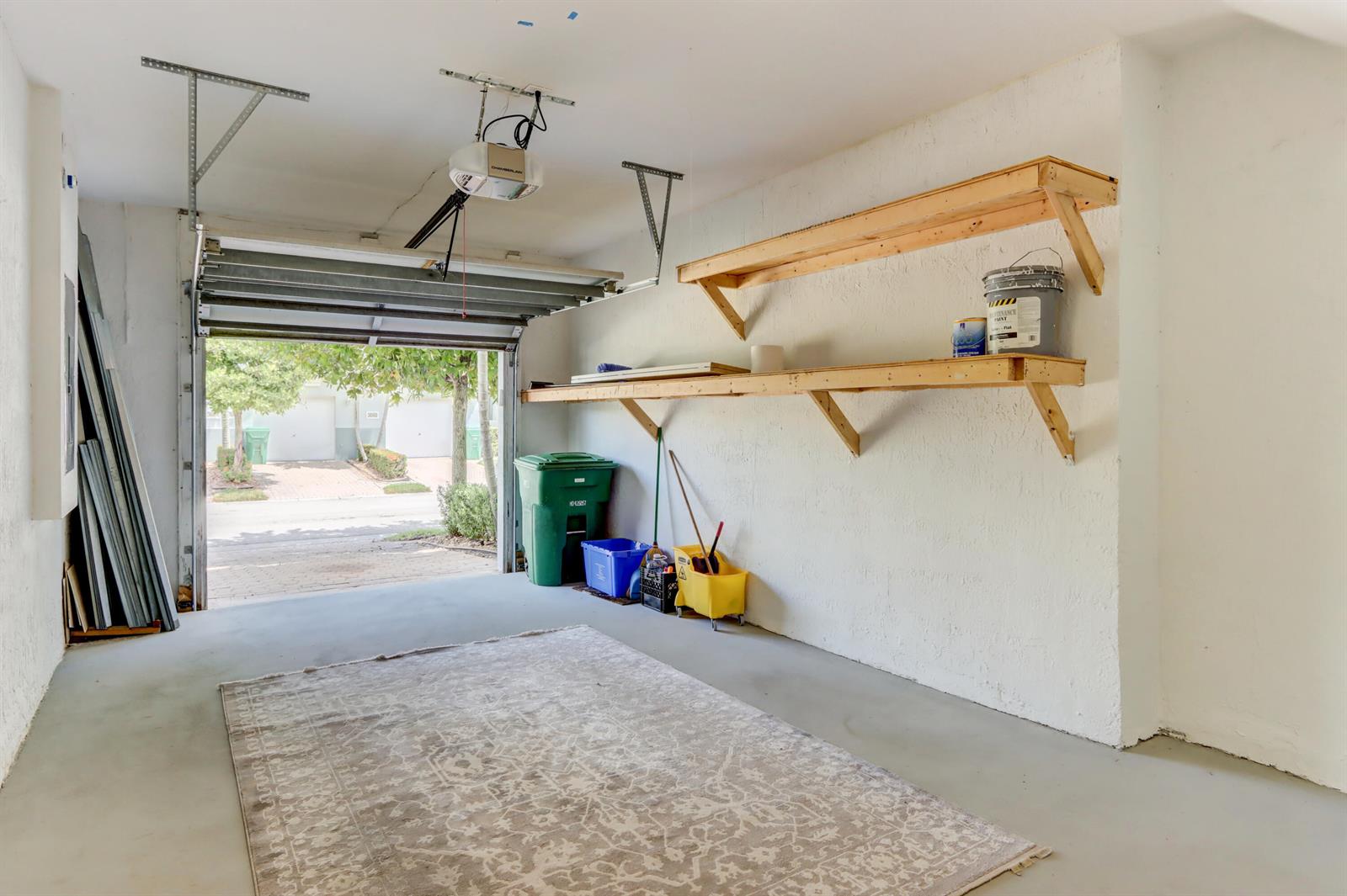 An open garage features a concrete floor, a large area rug, and wooden wall-mounted shelves — 3061 Laurel Ridge Circle, Riviera Beach FL 33404, RIVIERA BEACH, FL