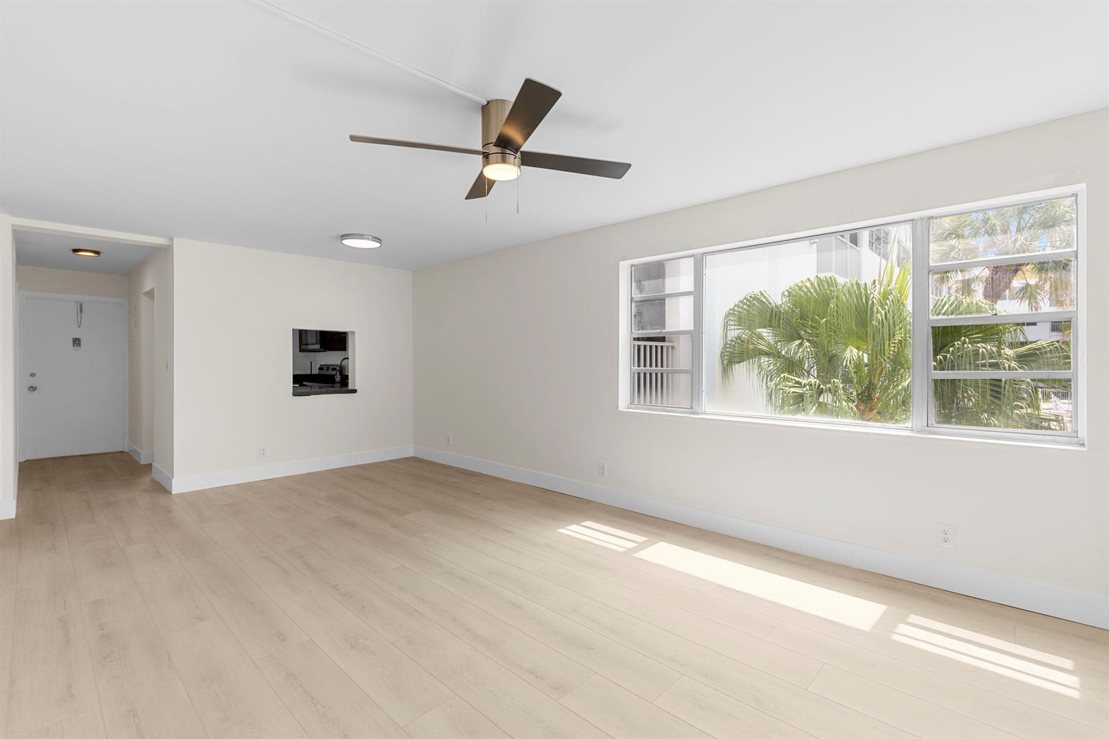 An empty living room features light wood-look flooring, a ceiling fan, and a large window — 3201 NE 8th Street # 206, Pompano Beach FL 33062, POMPANO BEACH, FL
