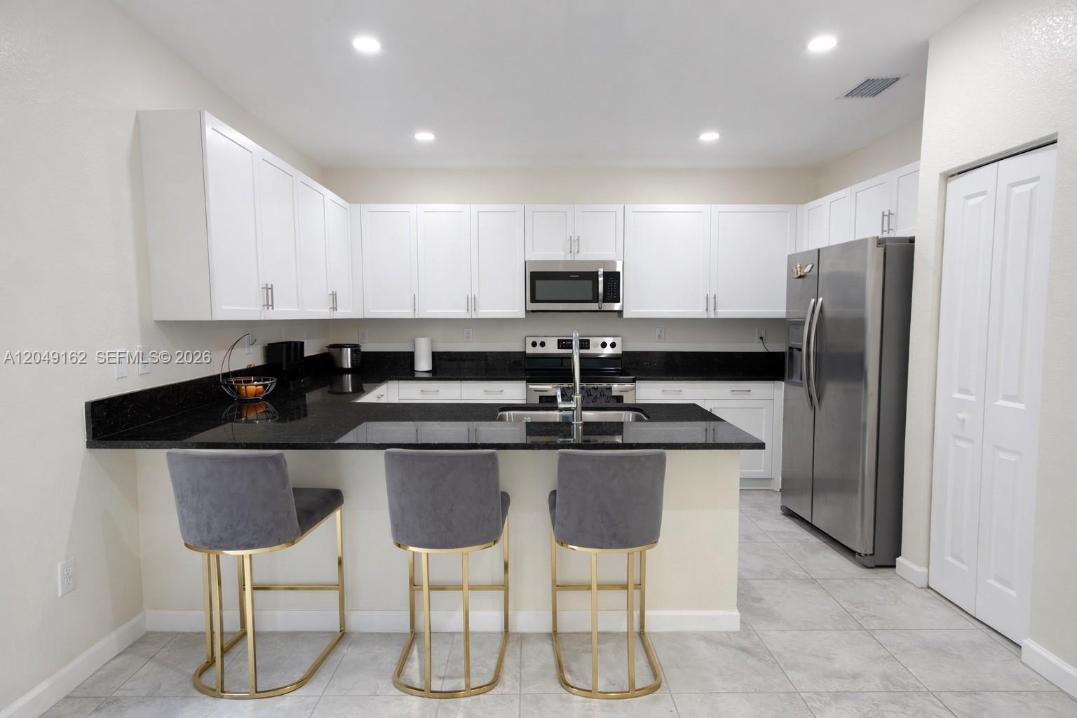 A kitchen features white cabinets, black countertops, stainless steel appliances, and three gray bar stools — 323 NE 208th Ter # 0, Miami FL 33179, MIAMI, FL