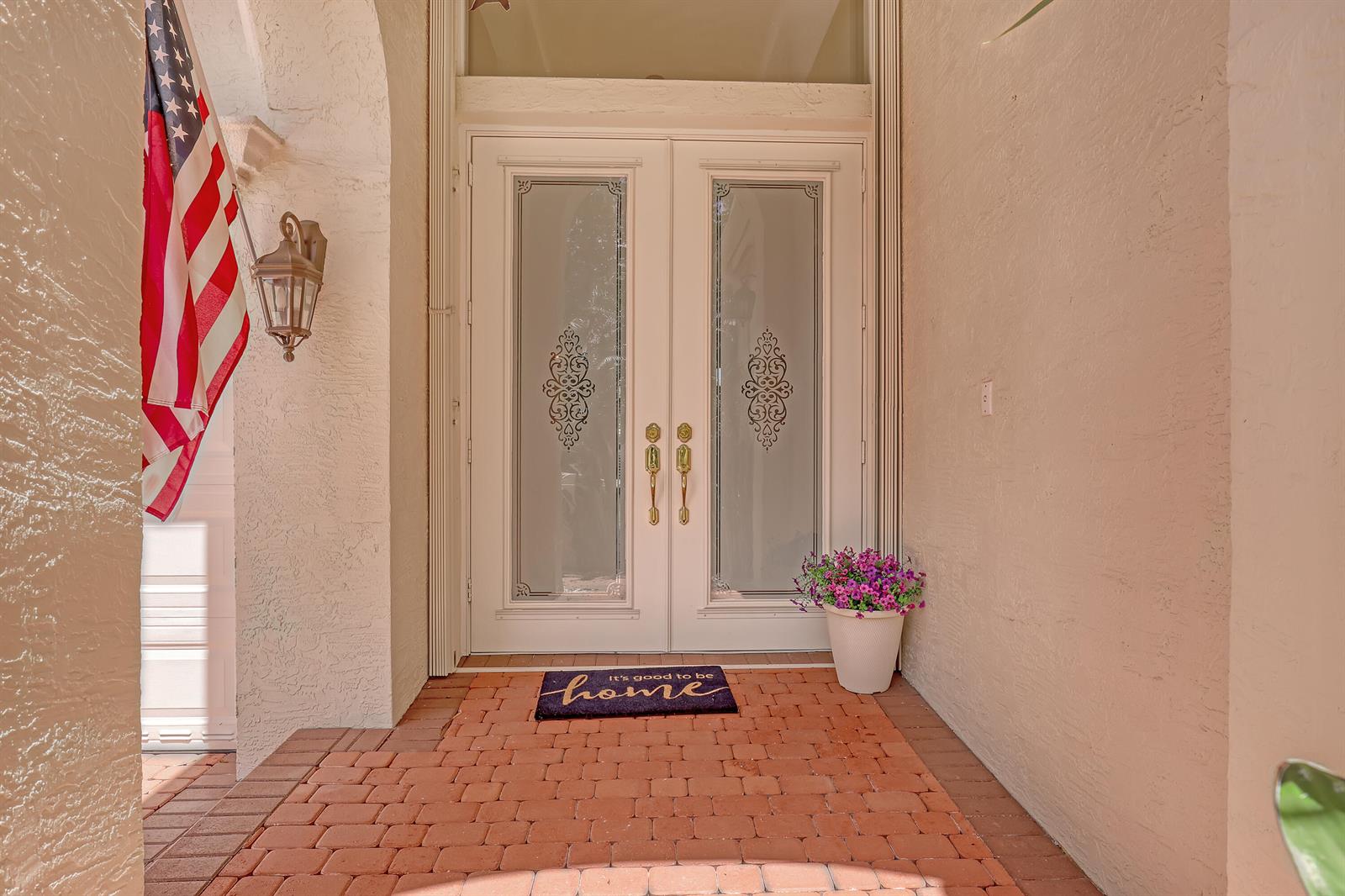 A brick-paved entryway features double glass-paneled doors, a potted plant, and an American flag — 9493 Via Elegante, Wellington FL 33411, WELLINGTON, FL