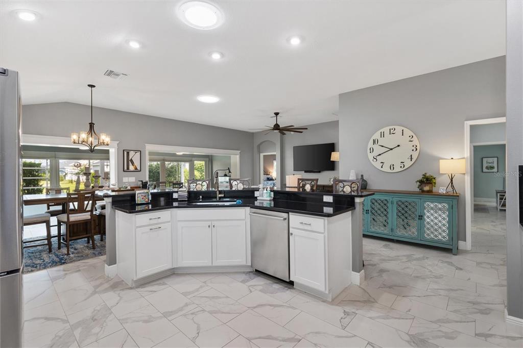 A kitchen with white cabinets, black countertops, and light-colored tile flooring opens to a dining area — 5852 ABNEY AVENUE, THE VILLAGES, FL