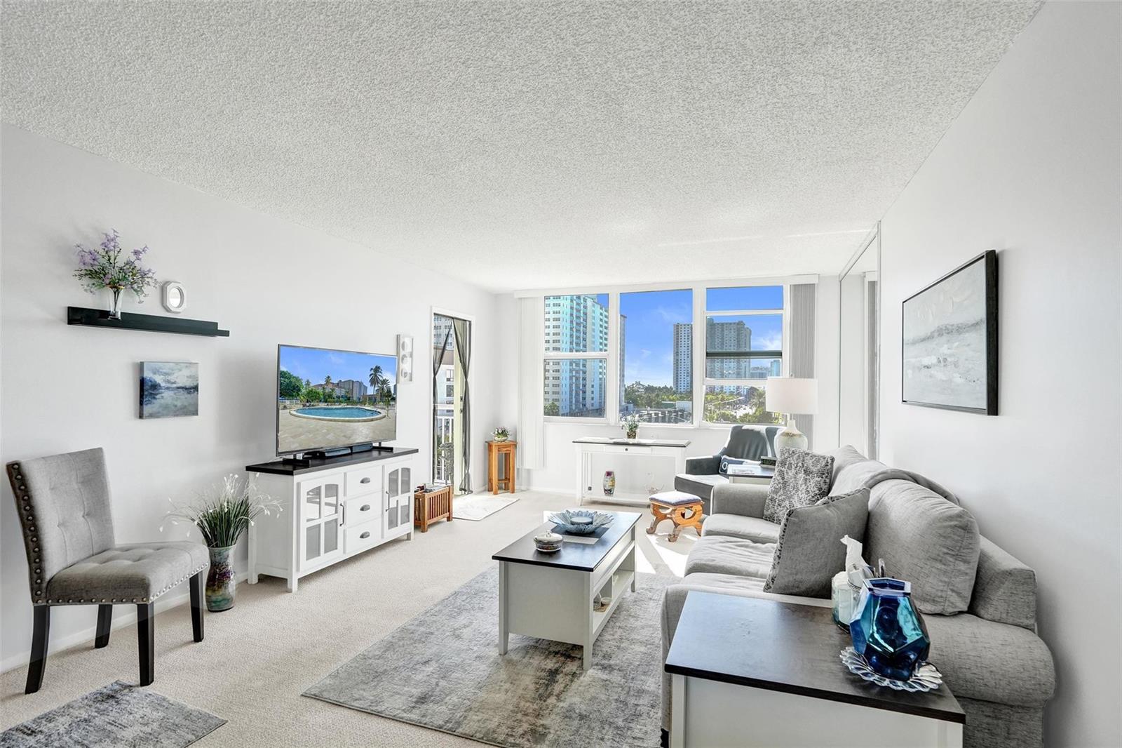 A living room features a gray sofa, white media console, and large windows with city views — 201 N Ocean Boulevard # 603, Pompano Beach FL 33062, POMPANO BEACH, FL
