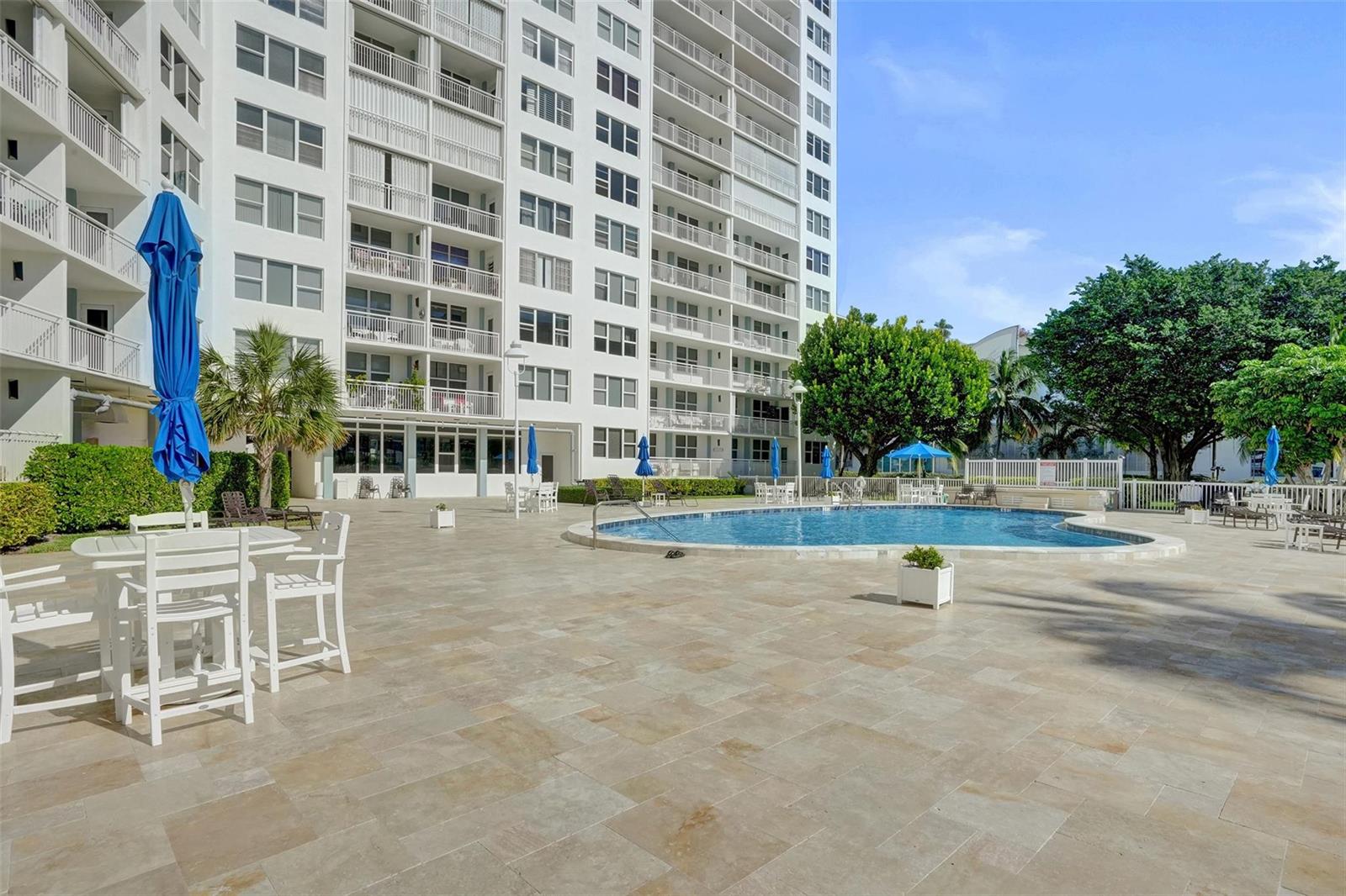 An outdoor pool area with a large apartment building and patio furniture under blue umbrellas — 201 N Ocean Boulevard # 603, Pompano Beach FL 33062, POMPANO BEACH, FL