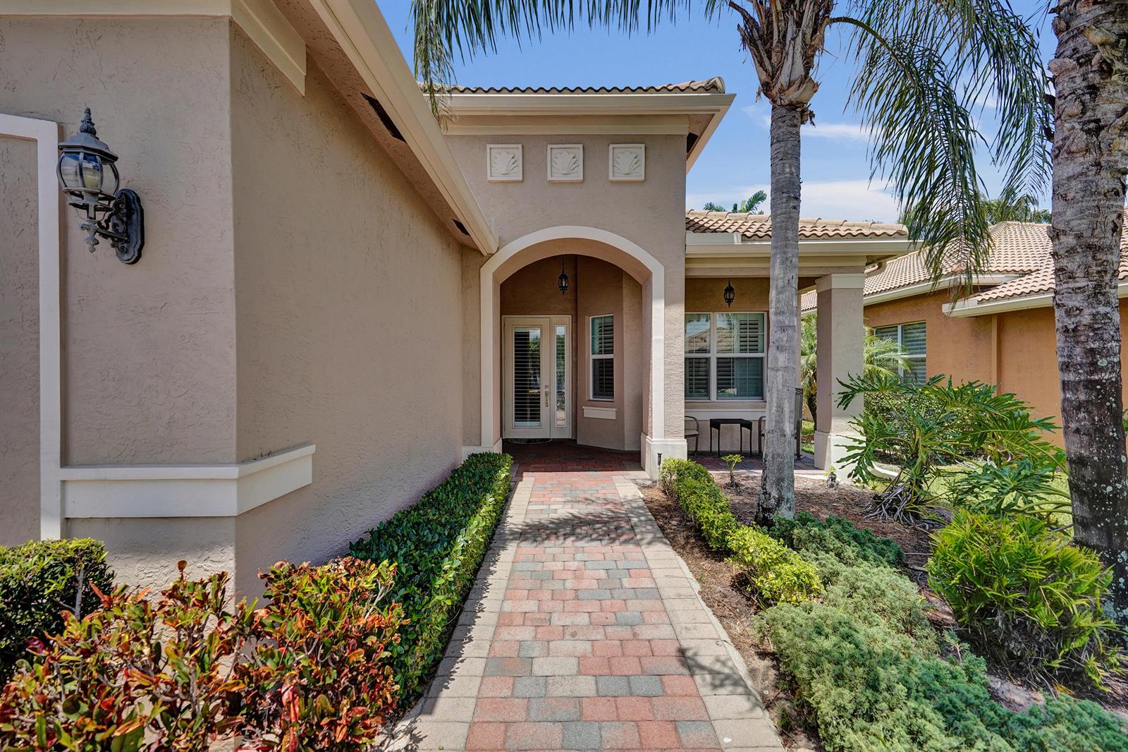 A brick walkway leads to the arched entryway of a tan stucco house with palm trees — 8703 Carmel Mountain Way, Boynton Beach FL 33473, BOYNTON BEACH, FL