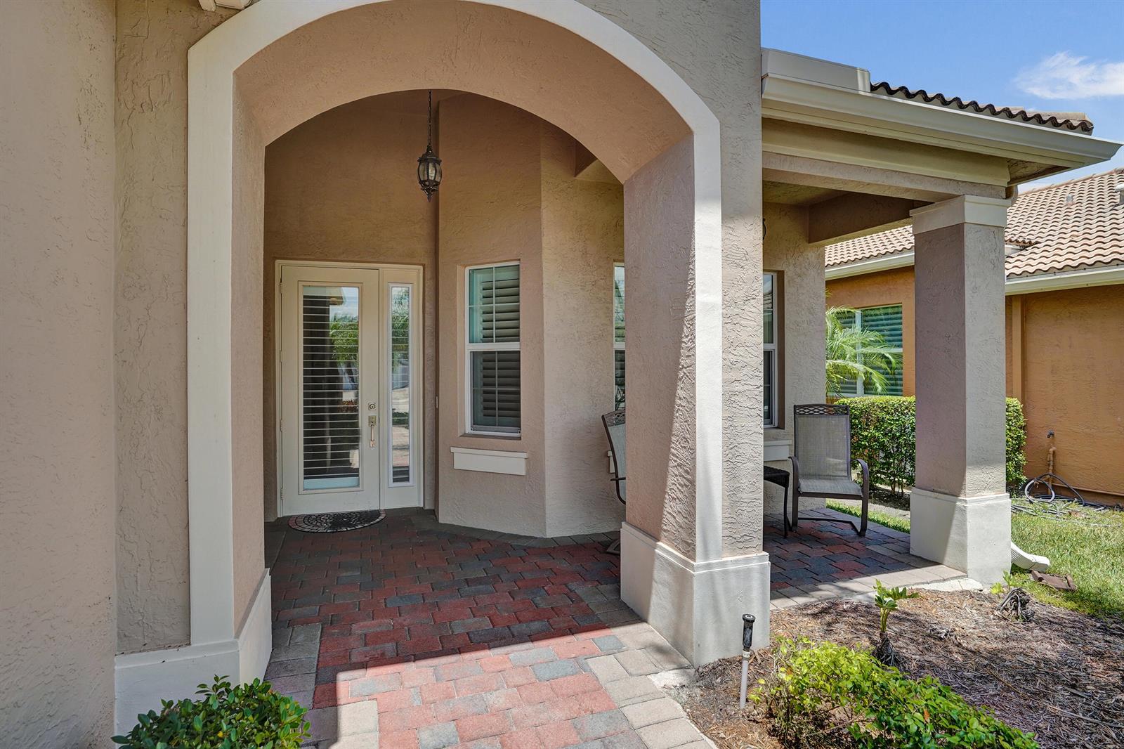 A covered entryway features a brick paver floor, a glass-paneled door, and beige stucco walls — 8703 Carmel Mountain Way, Boynton Beach FL 33473, BOYNTON BEACH, FL
