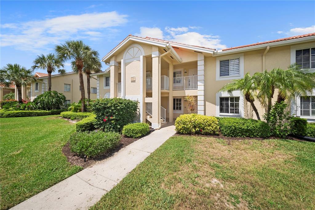 A two-story beige apartment building with a tiled roof, palm trees, and a concrete walkway — 7761 JEWEL LANE UNIT 102, NAPLES, FL