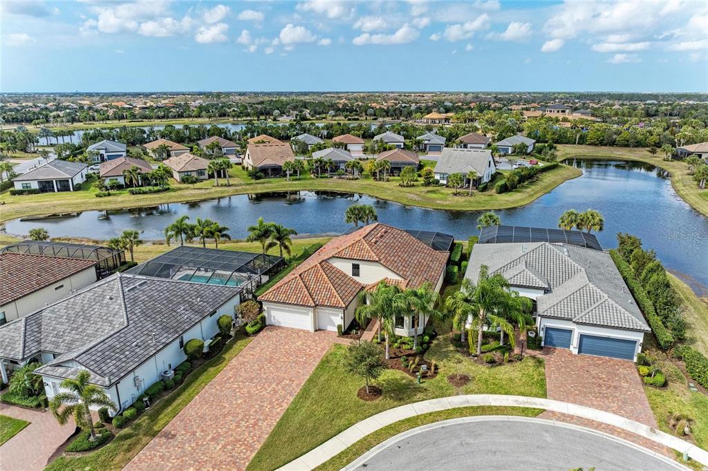 An aerial view shows three suburban houses with tiled roofs and driveways near a lake — 7110 GRADFORD COURT, LAKEWOOD RANCH, FL