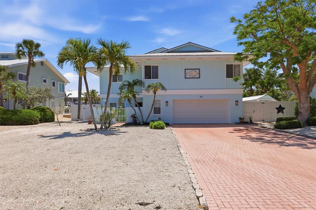 A two-story light blue house features a brick driveway, a garage, and palm trees — 517 KUMQUAT DRIVE, ANNA MARIA, FL