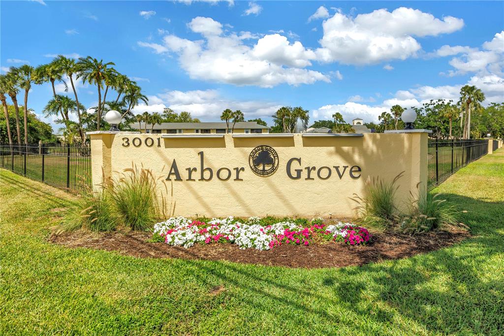 A beige stucco monument sign for Arbor Grove sits in a grassy yard with landscaping — 3001 58TH AVENUE S UNIT 315, ST PETERSBURG, FL