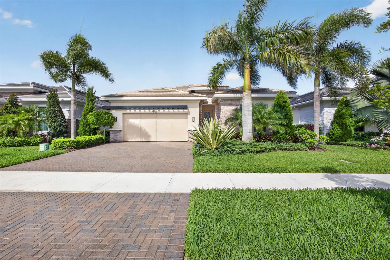 A single-story house with a beige garage, brick driveway, and palm trees in the front yard — 9941 Regency Way, Palm Beach Gardens FL 33412, PALM BEACH GARDENS, FL