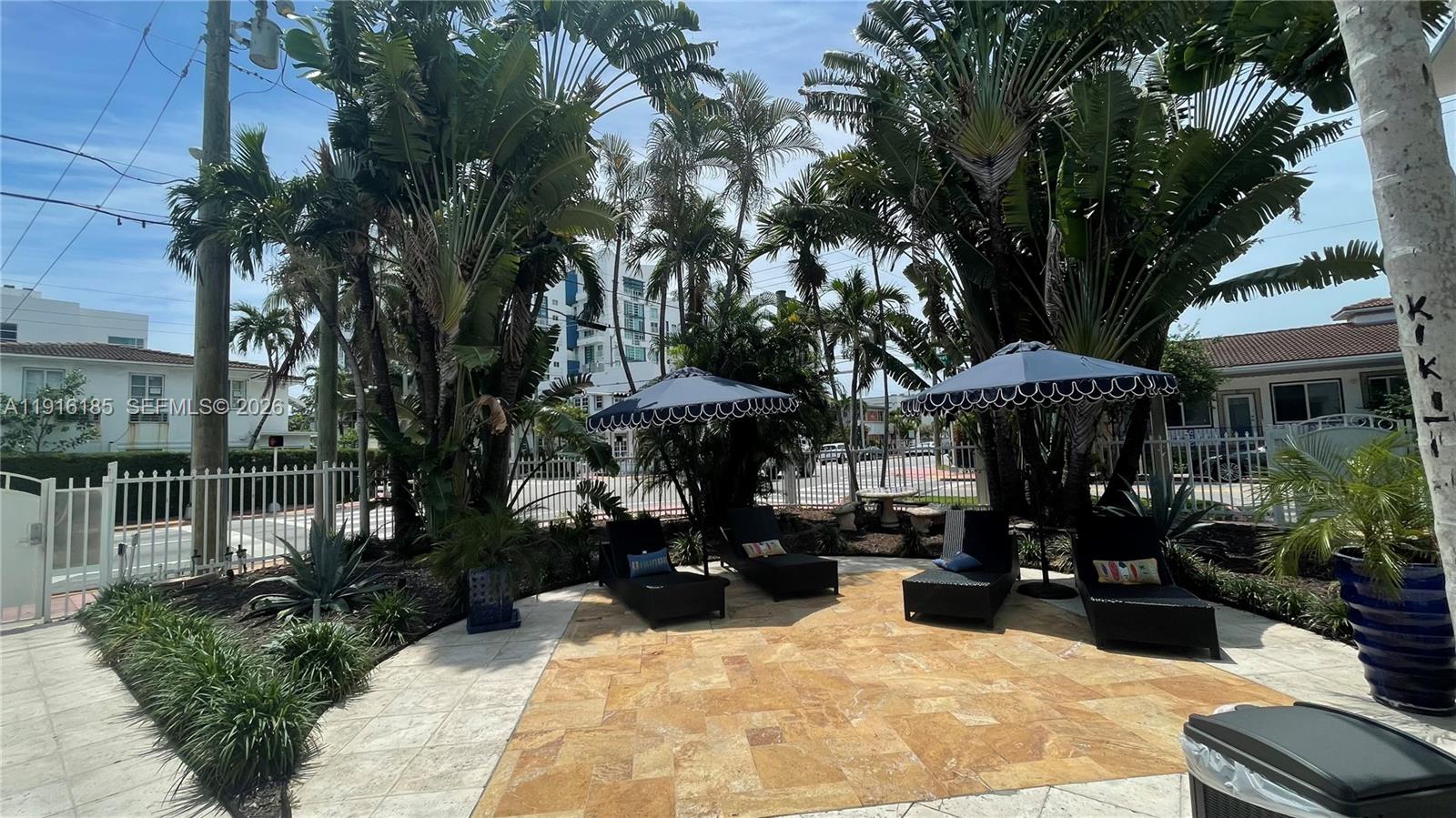 A stone patio features four lounge chairs under two umbrellas surrounded by tropical landscaping — 7710 Harding Ave # 408, Miami Beach FL 33141, MIAMI BEACH, FL