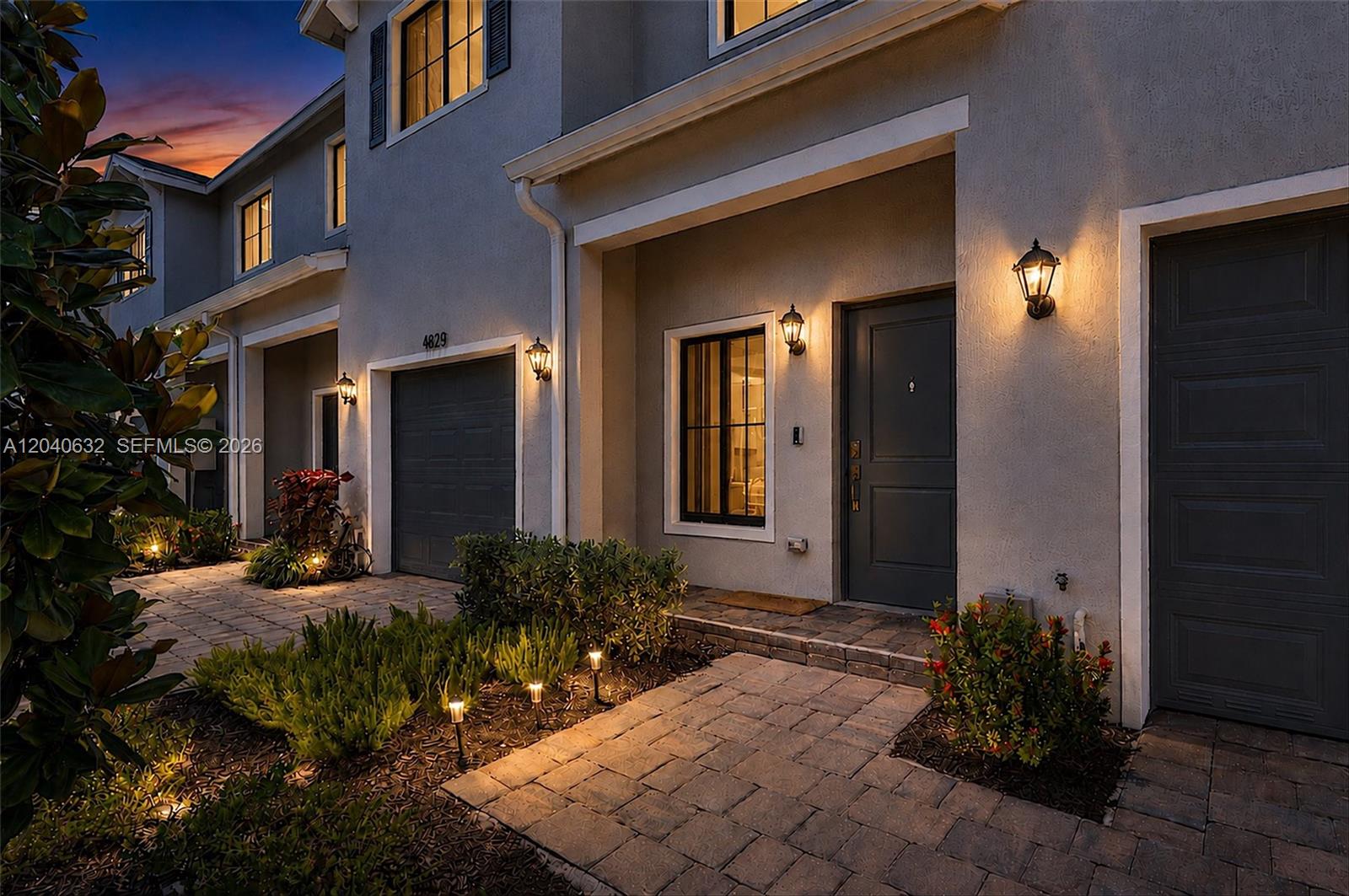A two-story townhouse exterior features a paved walkway, garage, and warm outdoor lighting at dusk — 4825 Dillion St, Lake Worth FL 33463, LAKE WORTH, FL