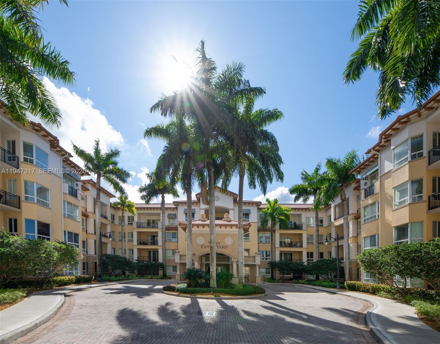 A paved circular driveway leads to the entrance of a multi-story apartment building with palms — 16101 Emerald Estates Dr # 250, Weston FL 33331, WESTON, FL