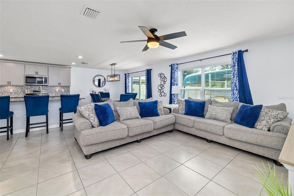 An open-concept living room and kitchen featuring gray sofas, blue accents, and tile flooring — 17315 WHEATBERRY BOULEVARD, PARRISH, FL