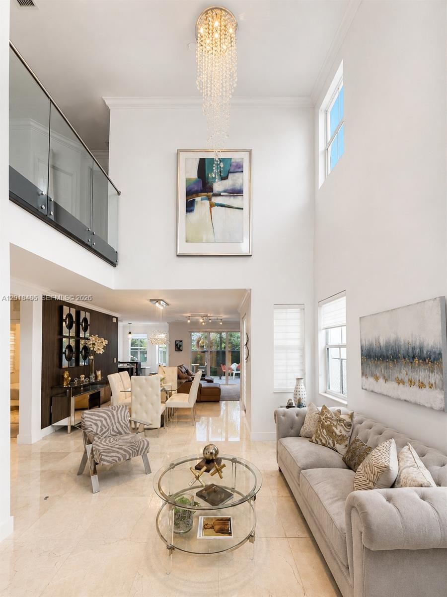 A high-ceilinged living room features a crystal chandelier, glass-railed balcony, and neutral tile flooring — 6950 NW 106th Ave, Doral FL 33178, DORAL, FL