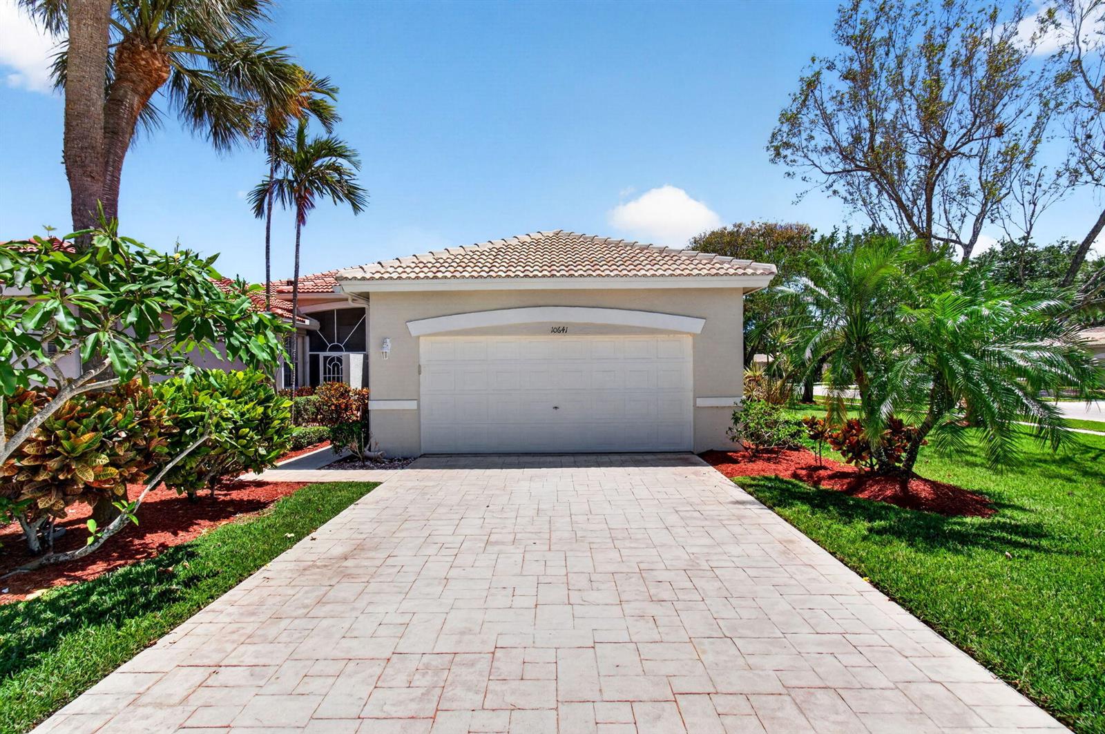 A beige house with a tiled roof and a paved driveway sits under a blue sky — 10641 Royal Caribbean Circle, Boynton Beach FL 33437, BOYNTON BEACH, FL