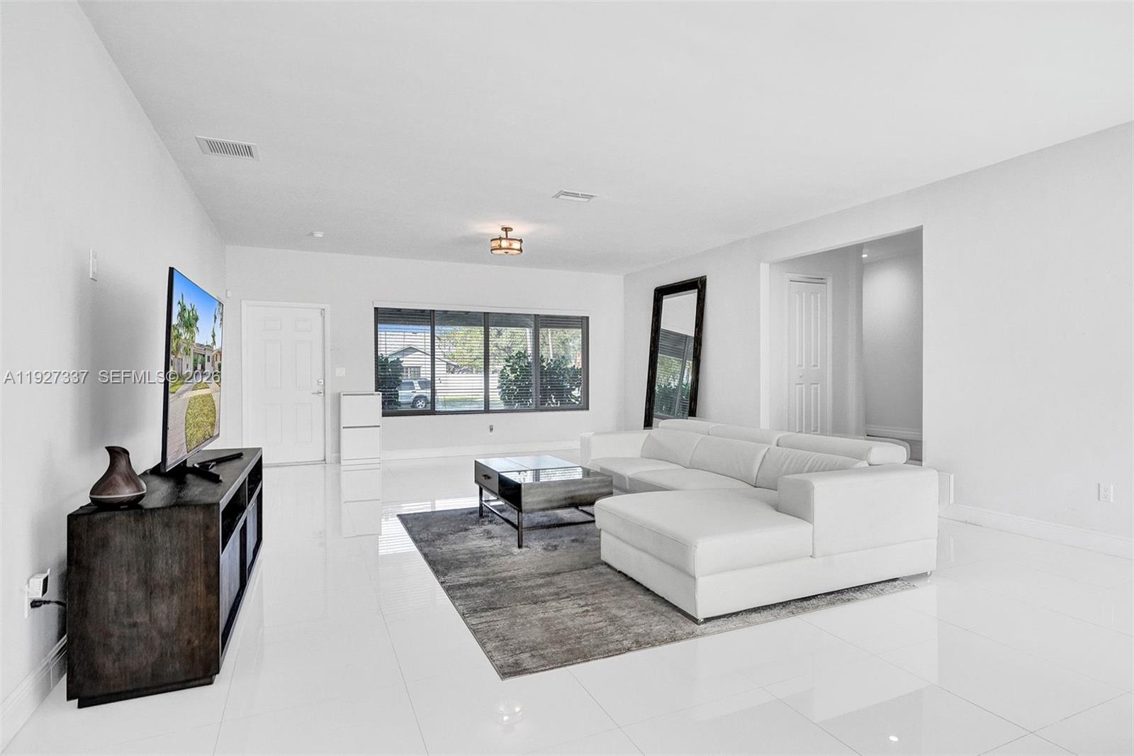 A bright living room features white tile floors, a white sectional sofa, and a television — 1102 Meadowlark Ave, Miami Springs FL 33166, MIAMI SPRINGS, FL