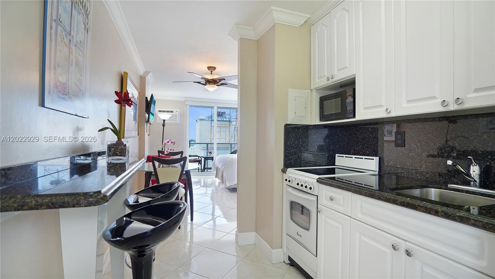 A kitchen with white cabinets and black countertops opens into a living area with balcony — 1750 James Ave # 9J, Miami Beach FL 33139, MIAMI BEACH, FL