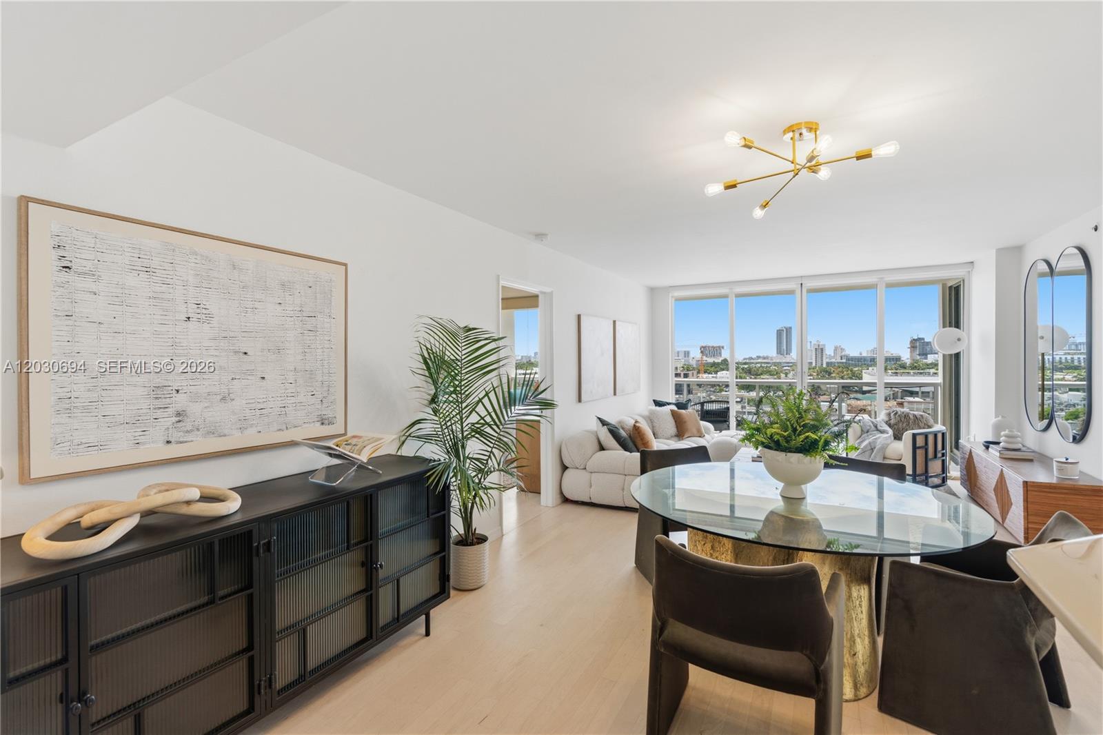 A bright living and dining area features a glass table, modern chandelier, and balcony view — 1800 Sunset Harbour Dr # 1003, Miami Beach FL 33139, MIAMI BEACH, FL