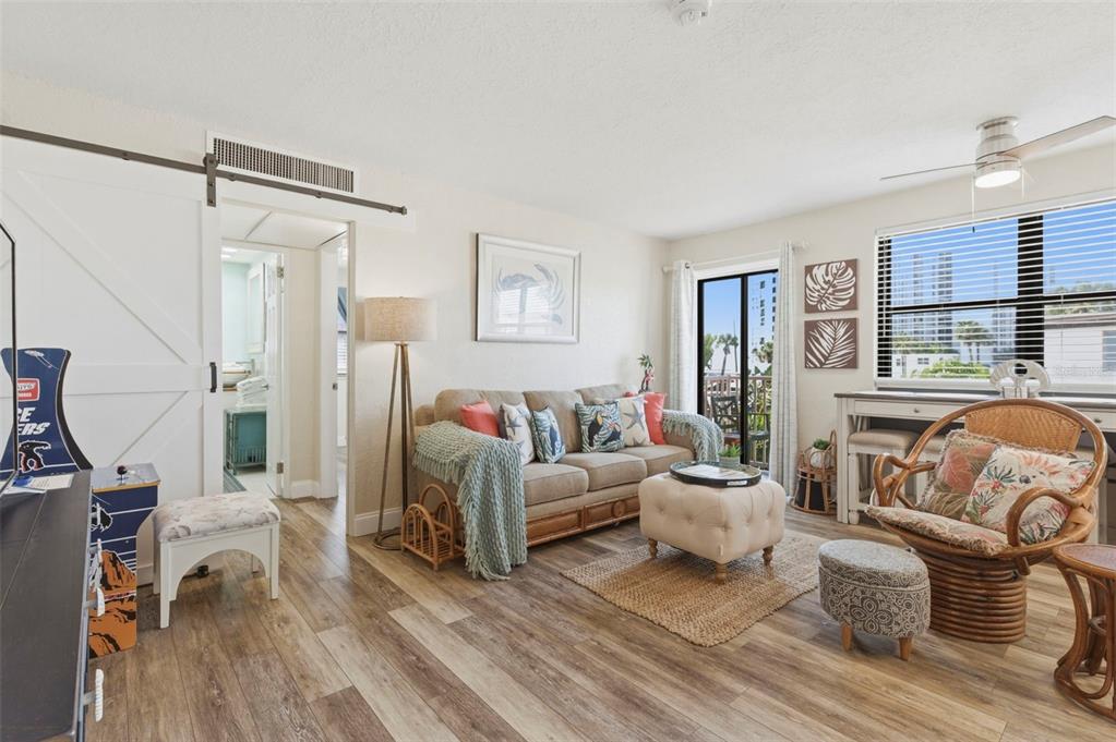 A living room features a beige sofa, wicker chair, sliding barn door, and wood-look flooring — 6800 SUNSET WAY UNIT 106, ST PETE BEACH, FL