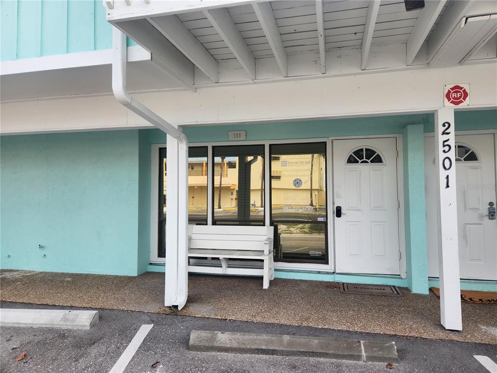 The exterior of a teal building features a white bench, a glass door, and a window — 2501 GULF DRIVE N UNIT 103, BRADENTON BEACH, FL