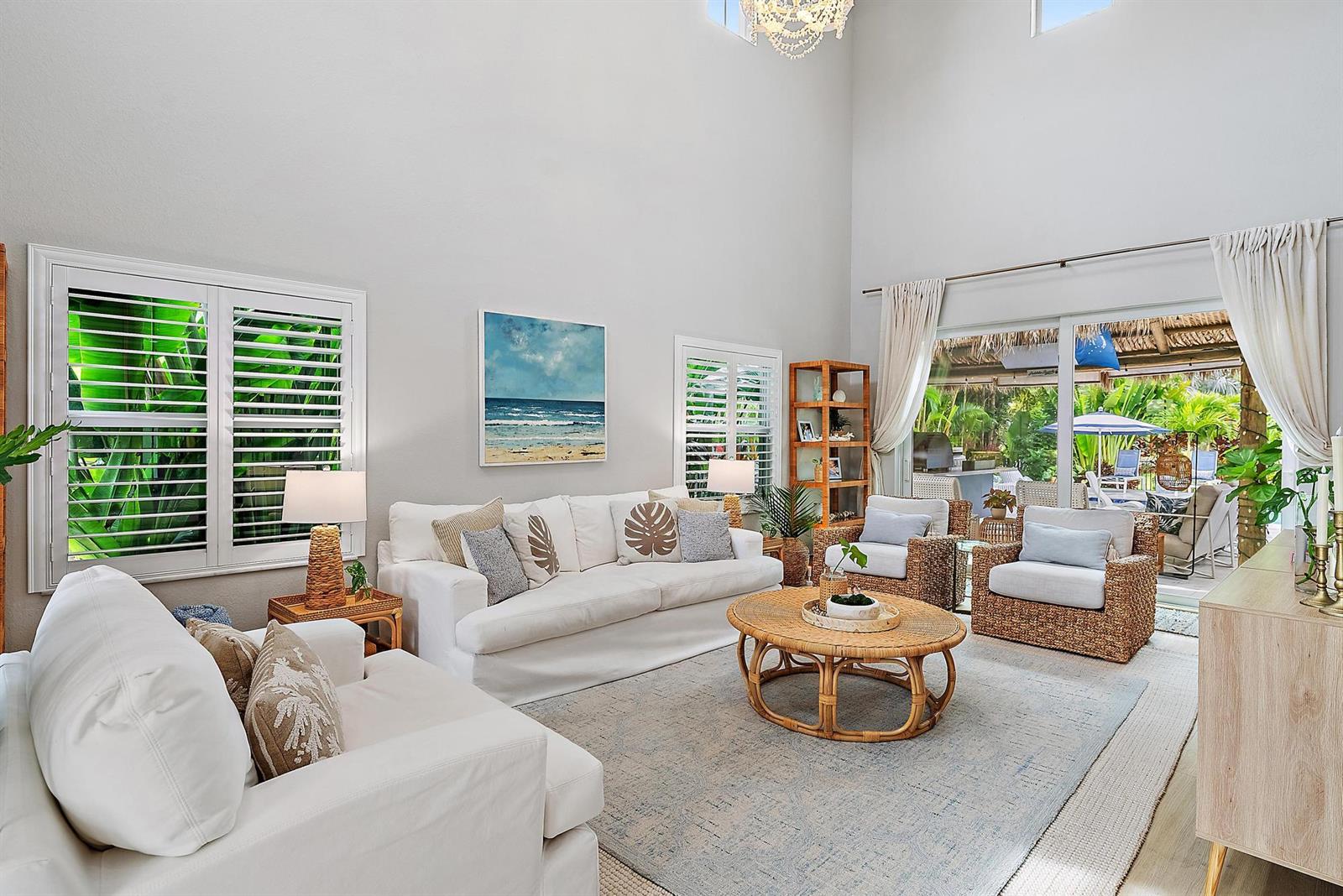 A bright living room features white sofas, rattan furniture, and large windows overlooking a patio — 152 Porgee Rock Place, Jupiter FL 33458, JUPITER, FL