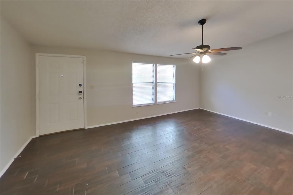 An empty living room features dark wood-look flooring, light walls, and a ceiling fan — 8102 CHICKASAW LANE, PORT RICHEY, FL