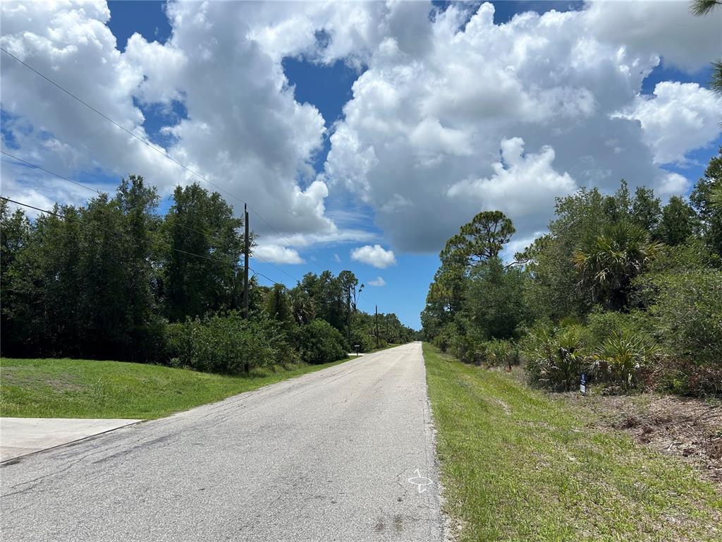 A paved road stretches into the distance between wooded areas under a cloudy blue sky — 17504 POSTON AVENUE, PORT CHARLOTTE, FL