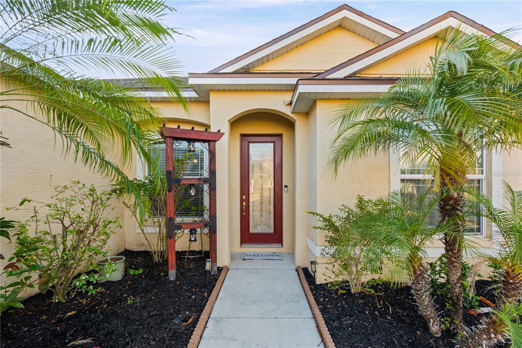 The front exterior of a yellow house features a red door and a wooden trellis — 12807 24TH STREET CIRCLE E, PARRISH, FL