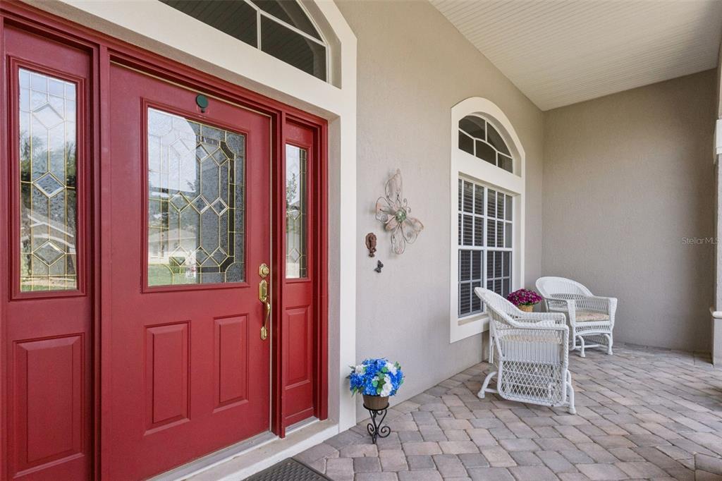 A red front door with glass panels sits on a paved porch with white chairs — 5607 SIR CHURCHILL DRIVE, LEESBURG, FL