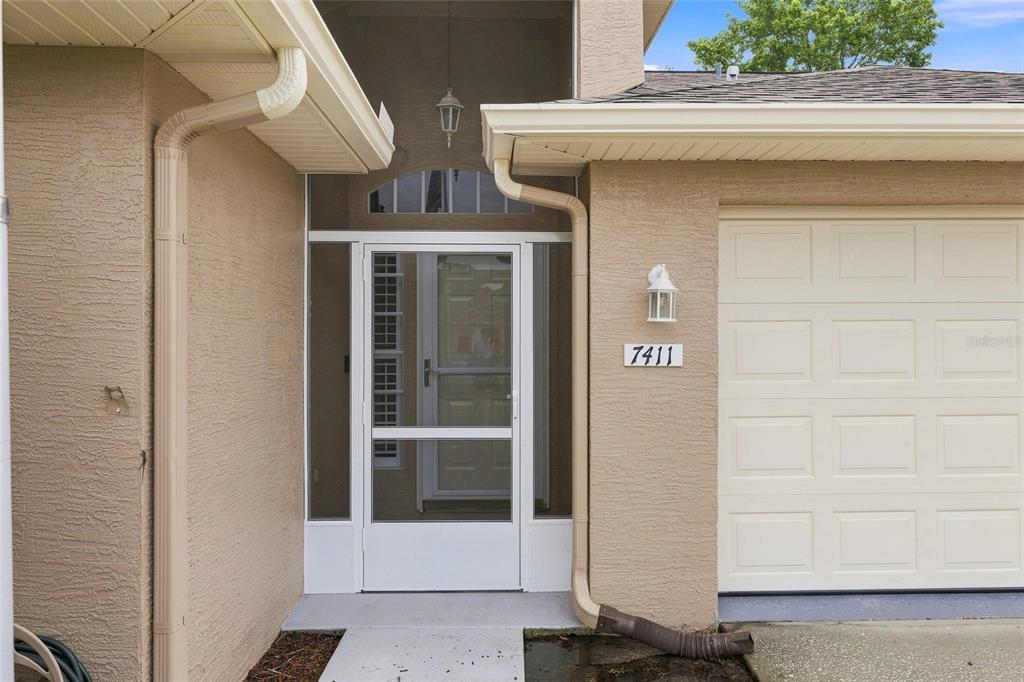 The exterior of a house features a tan stucco facade, a garage, and a screened entryway — 7411 MOORGATE COURT, NEW PORT RICHEY, FL