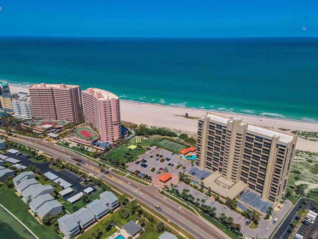 An aerial view shows high-rise beachfront condominiums, tennis courts, and a parking lot near the ocean — 1290 GULF BOULEVARD UNIT 2003, CLEARWATER BEACH, FL