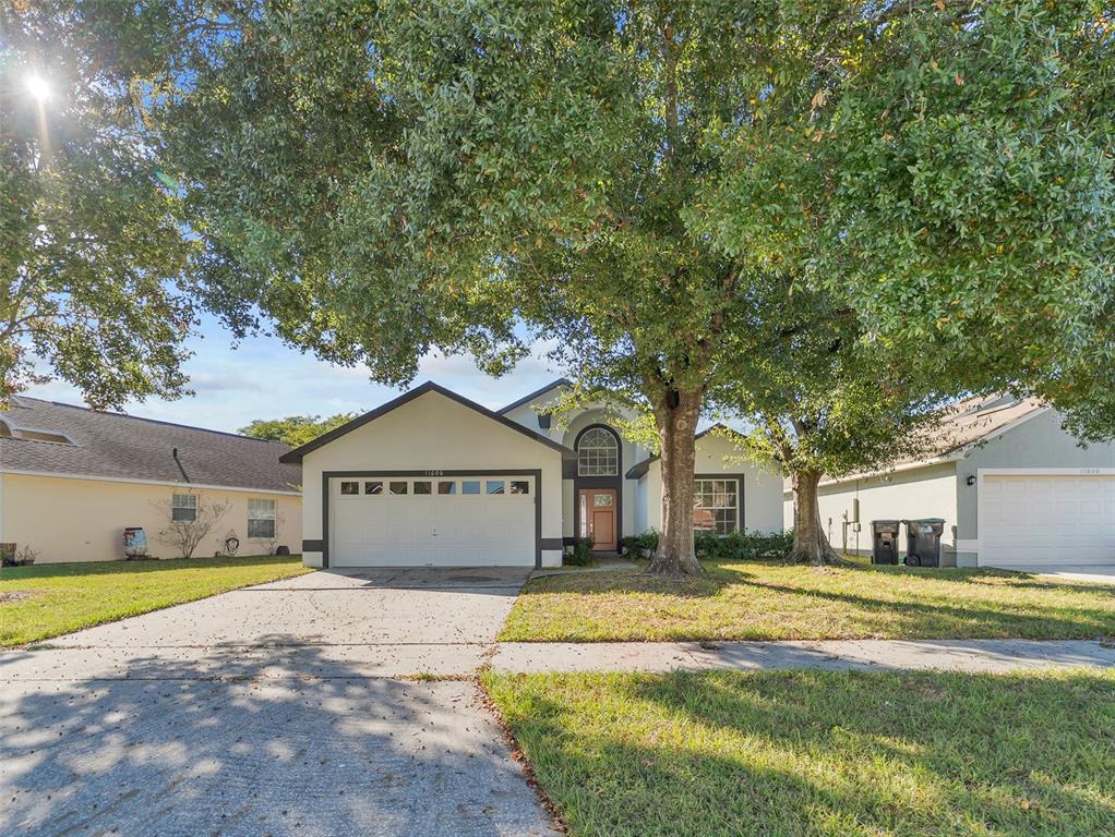 A single-story house with a two-car garage and large trees in the front yard — 11606 GOODWYCK DRIVE, ORLANDO, FL